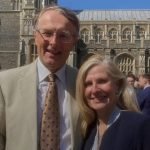 A man in a light suit and a woman in a dark jacket smile at the camera, with a church in the background