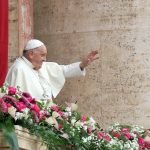 Pope Franics dressed in white robes waving to crowds from a balcony covered in pink flowers