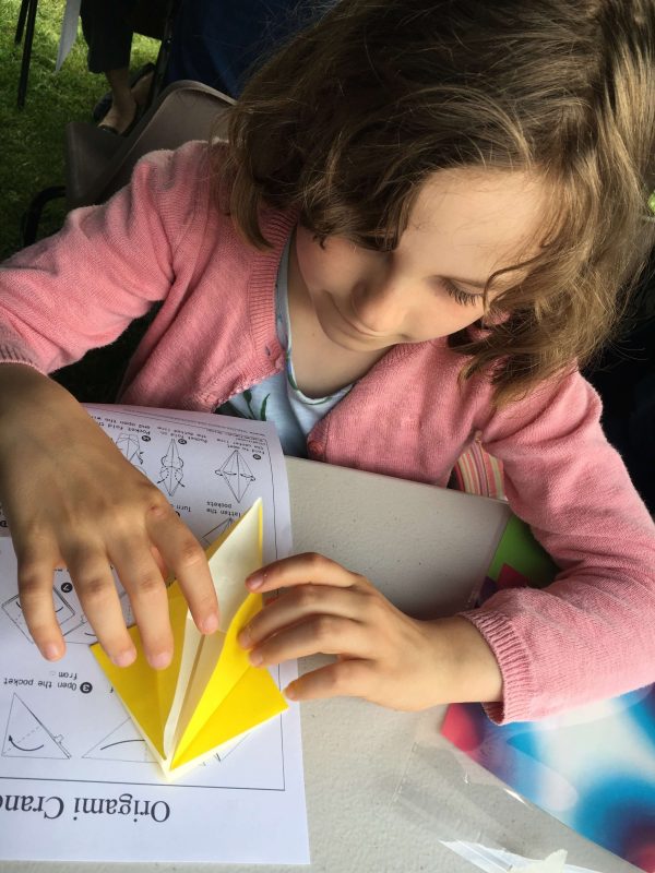 View of girl sitting at a table folding yellow origami paper to make a shape.