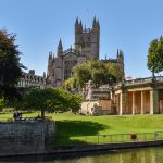 A city church with blue sky, trees with green leaves and green grass leading to a river in the foreground.